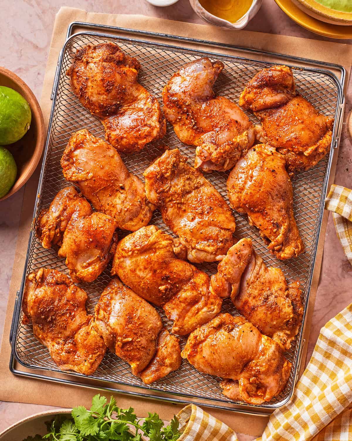 Seasoned raw chicken thighs arranged on a metal rack, ready for cooking. Fresh limes, cilantro, a yellow-checked napkin, and bowls are visible around the rack on a light-colored surface.