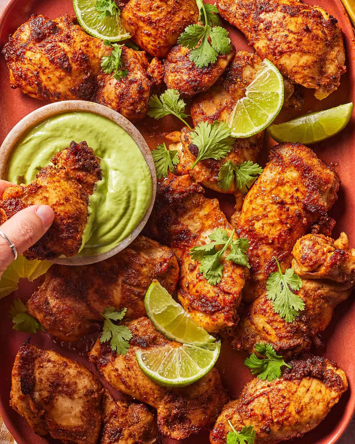 A platter of grilled chicken garnished with cilantro and lime wedges, with a hand dipping a piece into a bowl of creamy green sauce.
