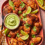 A platter of seasoned chicken pieces garnished with cilantro and lime wedges, served with a bowl of green dipping sauce and a fork on the side.