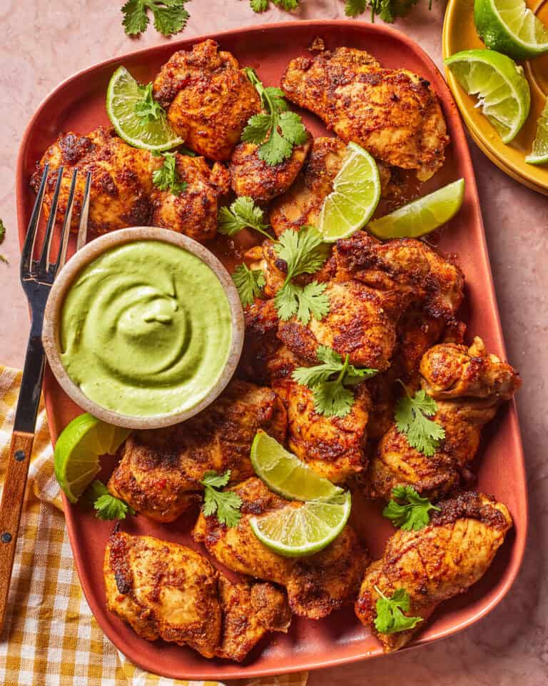 A platter of seasoned chicken pieces garnished with cilantro and lime wedges, served with a bowl of green dipping sauce and a fork on the side.