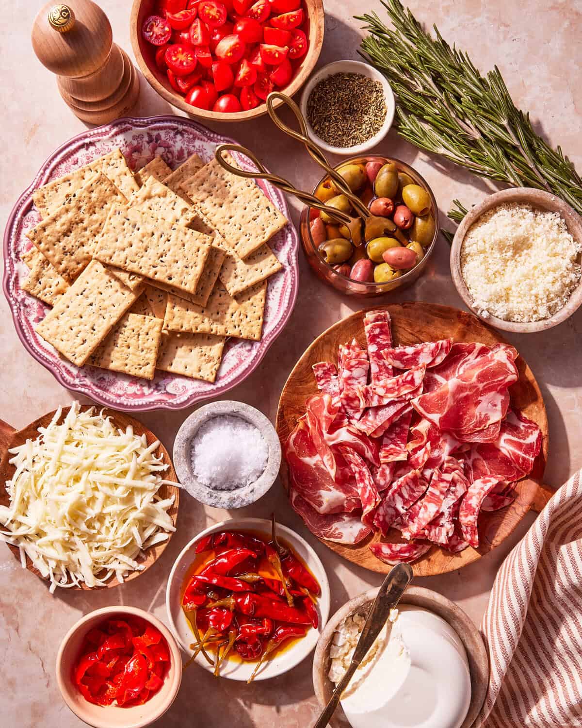 A charcuterie board with crackers, sliced cured meats, shredded cheese, olives, cherry tomatoes, peppers, grated cheese, herbs, spices, and salt, arranged on a light surface with a striped cloth.