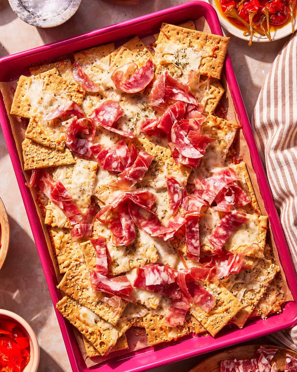 A pink tray filled with crackers topped with melted cheese, herbs, and slices of cured meat, surrounded by small bowls of cherries and a striped napkin.