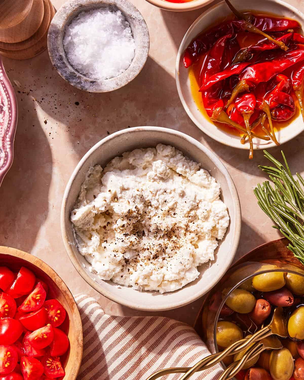 A bowl of ricotta cheese with cracked pepper surrounded by tomatoes, olives, marinated red peppers, rosemary, and a small dish of sea salt on a beige surface.