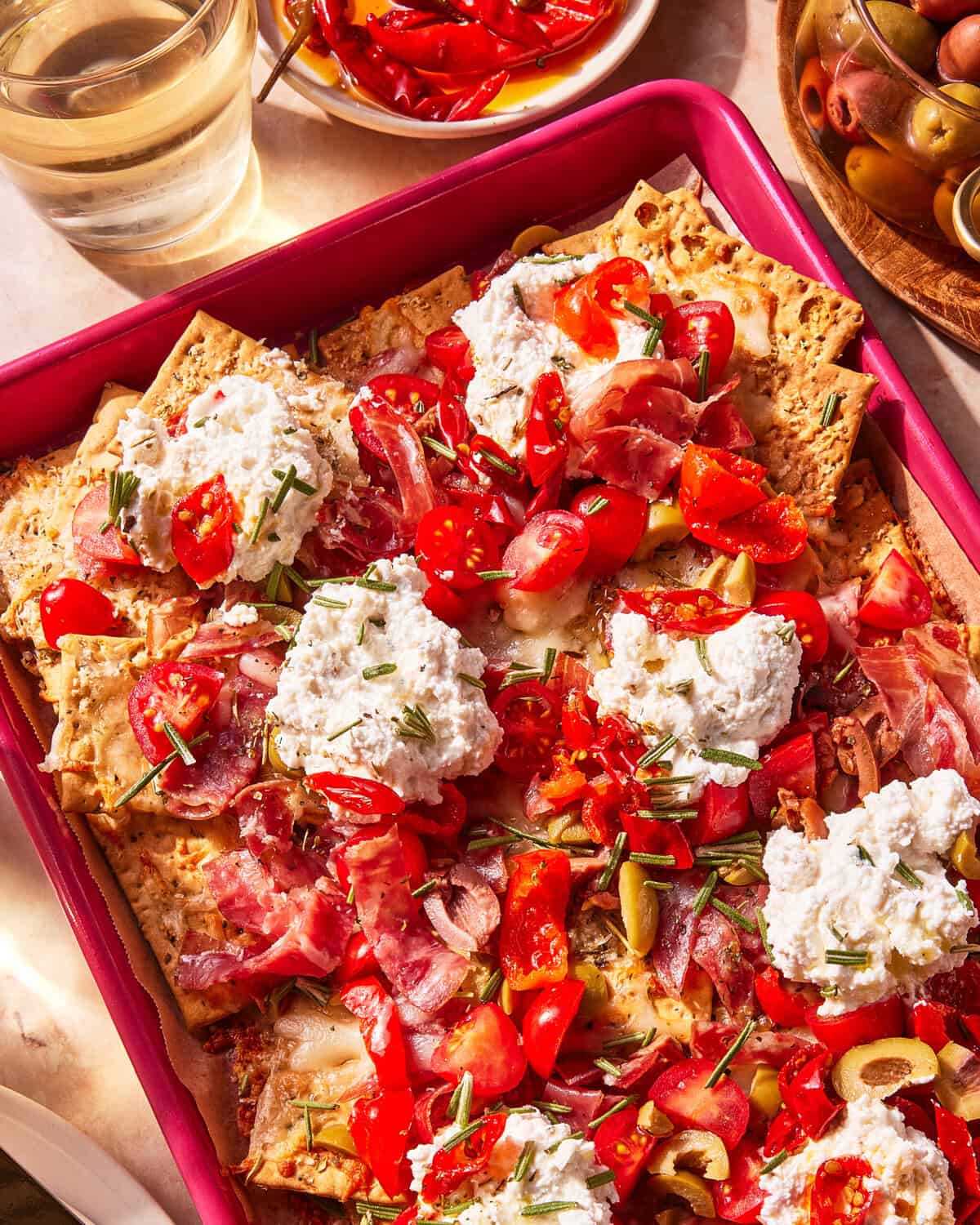 A tray of crispbread topped with dollops of ricotta, chopped tomatoes, ham, and herbs. Surrounding the tray are sliced red peppers, olives, and a glass of water. Sunlight brightens the colorful spread.