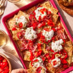 A tray of cracker nachos topped with ricotta cheese, sliced tomatoes, prosciutto, green olives, and herbs. Glasses of white wine, a bowl of olives, and more tomatoes are nearby. A hand reaches for a cracker.