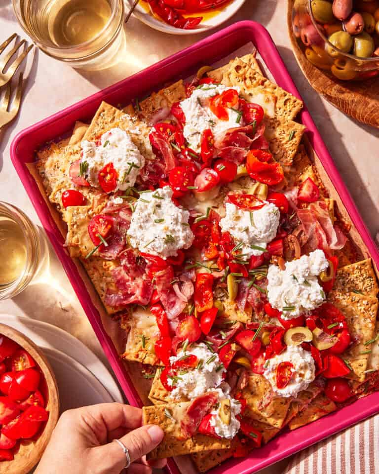 A tray of cracker nachos topped with ricotta cheese, sliced tomatoes, prosciutto, green olives, and herbs. Glasses of white wine, a bowl of olives, and more tomatoes are nearby. A hand reaches for a cracker.