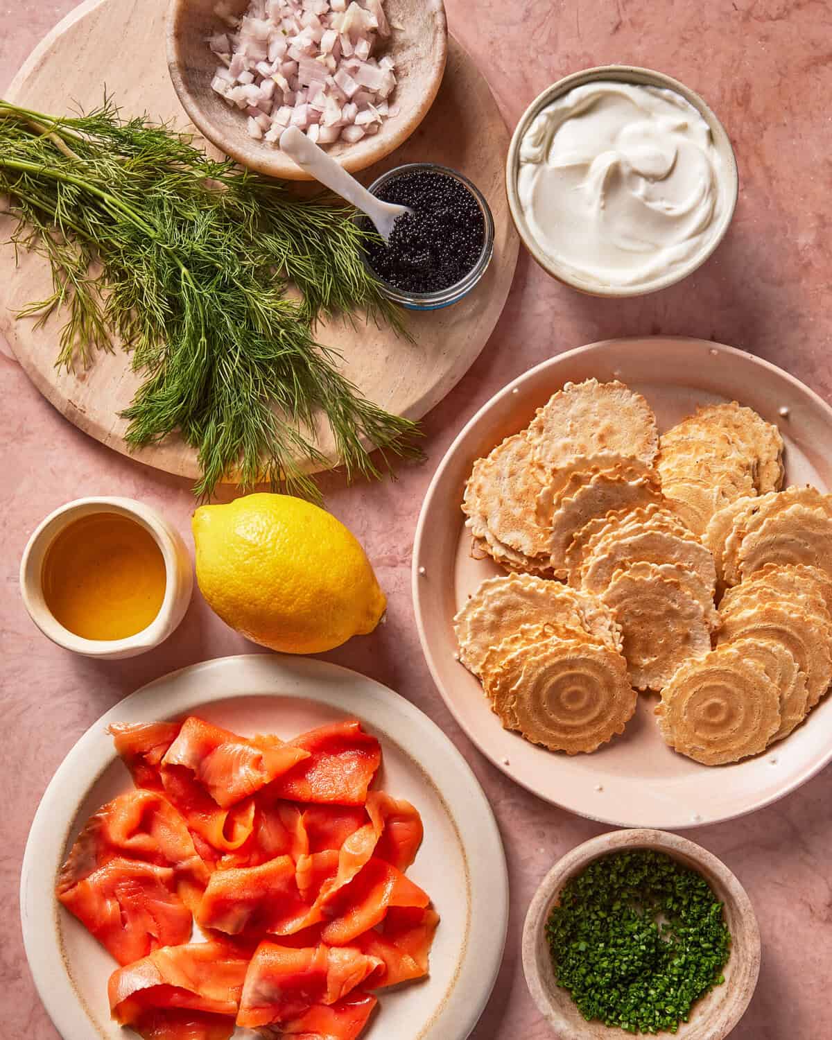 Crispy crackers, smoked salmon, lemon, fresh dill, chopped shallots, black caviar, sour cream, chives, and olive oil are arranged on pink marble. Ingredients are in bowls and plates, viewed from above.