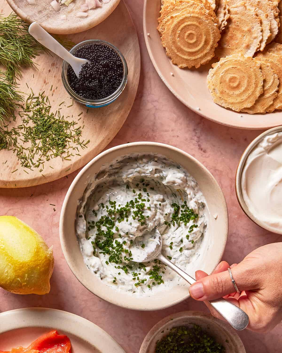 A hand stirs a bowl of creamy dip topped with chopped chives. Surrounding the bowl are crackers, herbs, caviar, lemon, and a dollop of sour cream on a pink surface.