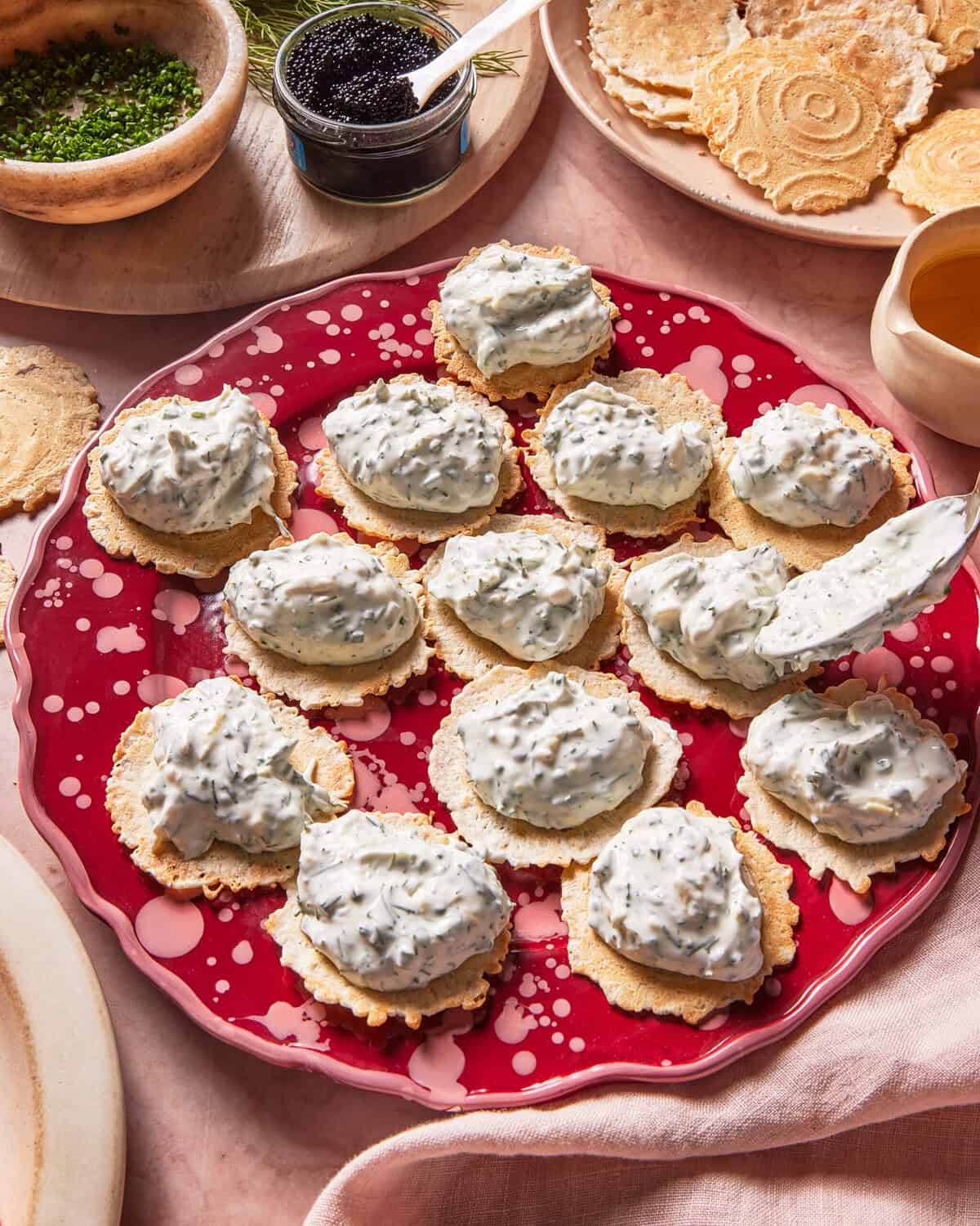 A red plate holds crisp crackers topped with a creamy herb spread. Nearby are bowls of chopped chives, black caviar, and a honey dipper, suggesting options for garnishing the appetizers.