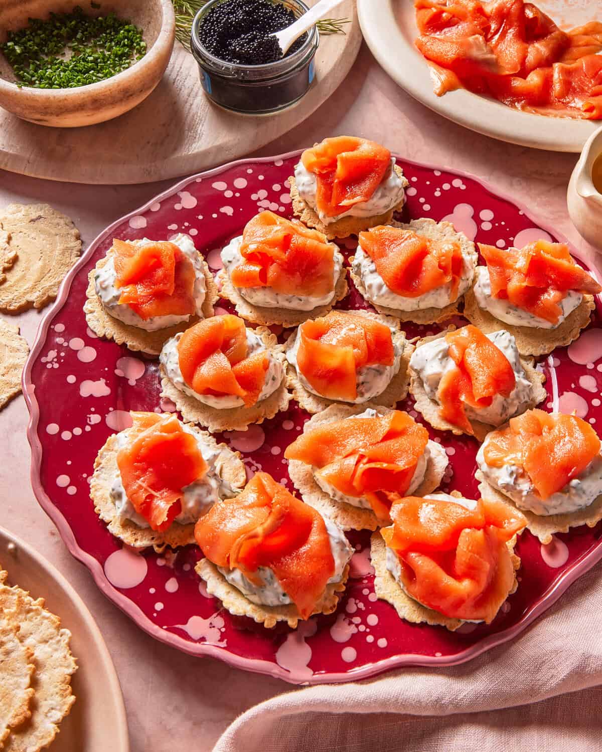 A red plate holds crackers topped with cream cheese and smoked salmon. Surrounding dishes contain chives, caviar, more crackers, and smoked salmon slices.