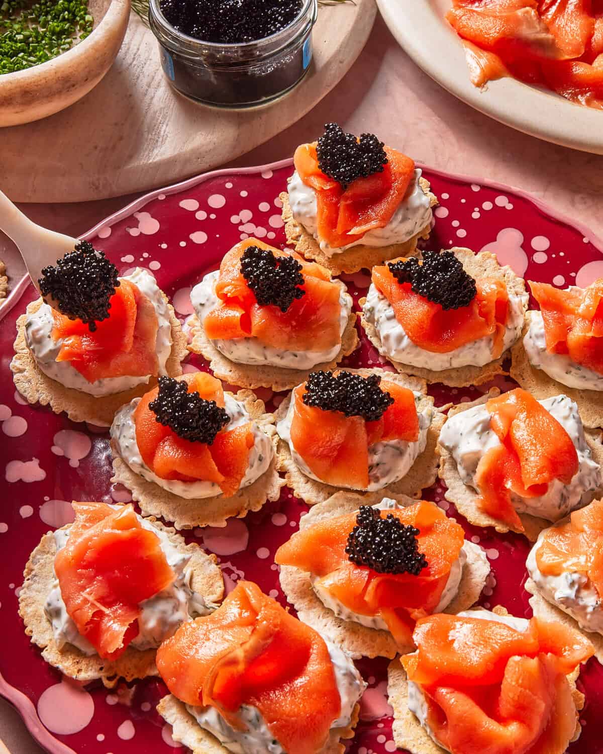 A red plate with crackers topped with creamy spread, smoked salmon, and a dollop of black caviar. A small jar of caviar and chopped herbs are nearby.