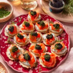 A red plate holds crackers topped with creamy spread, smoked salmon, black caviar, and fresh dill. Glasses of white wine, a jar of caviar, herbs, and stacked plates surround the platter on a pink surface.