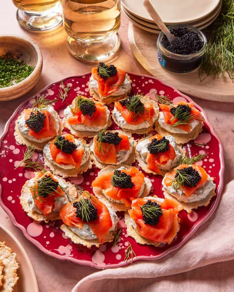 A red plate holds crackers topped with creamy spread, smoked salmon, black caviar, and fresh dill. Glasses of white wine, a jar of caviar, herbs, and stacked plates surround the platter on a pink surface.