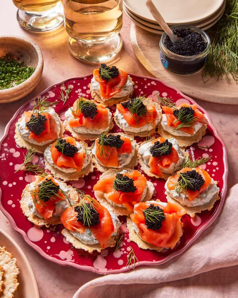 A red plate holds crackers topped with creamy spread, smoked salmon, black caviar, and fresh dill. Glasses of white wine, a jar of caviar, herbs, and stacked plates surround the platter on a pink surface.