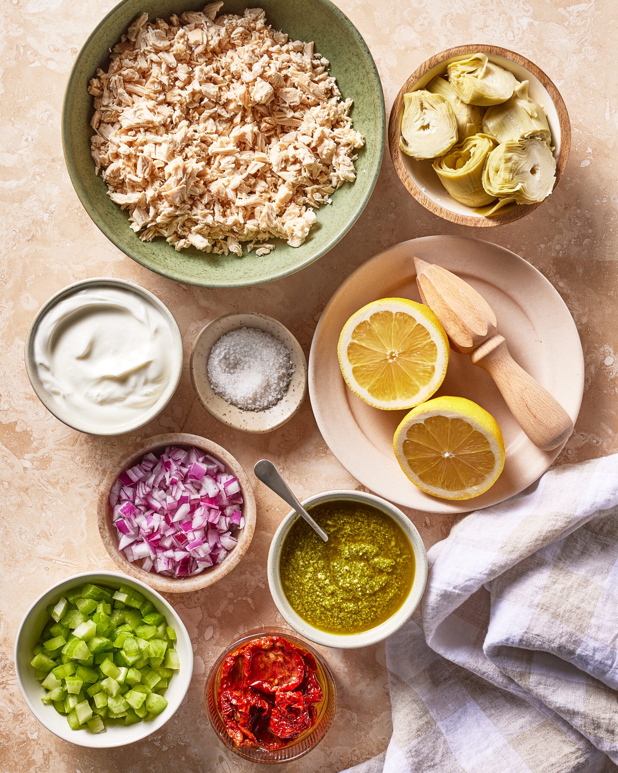 Bowls of shredded chicken, artichoke hearts, yogurt, chopped red onion, celery, sun-dried tomatoes, pesto, salt, and halved lemons with a wooden juicer on a beige surface next to a white and gray towel.