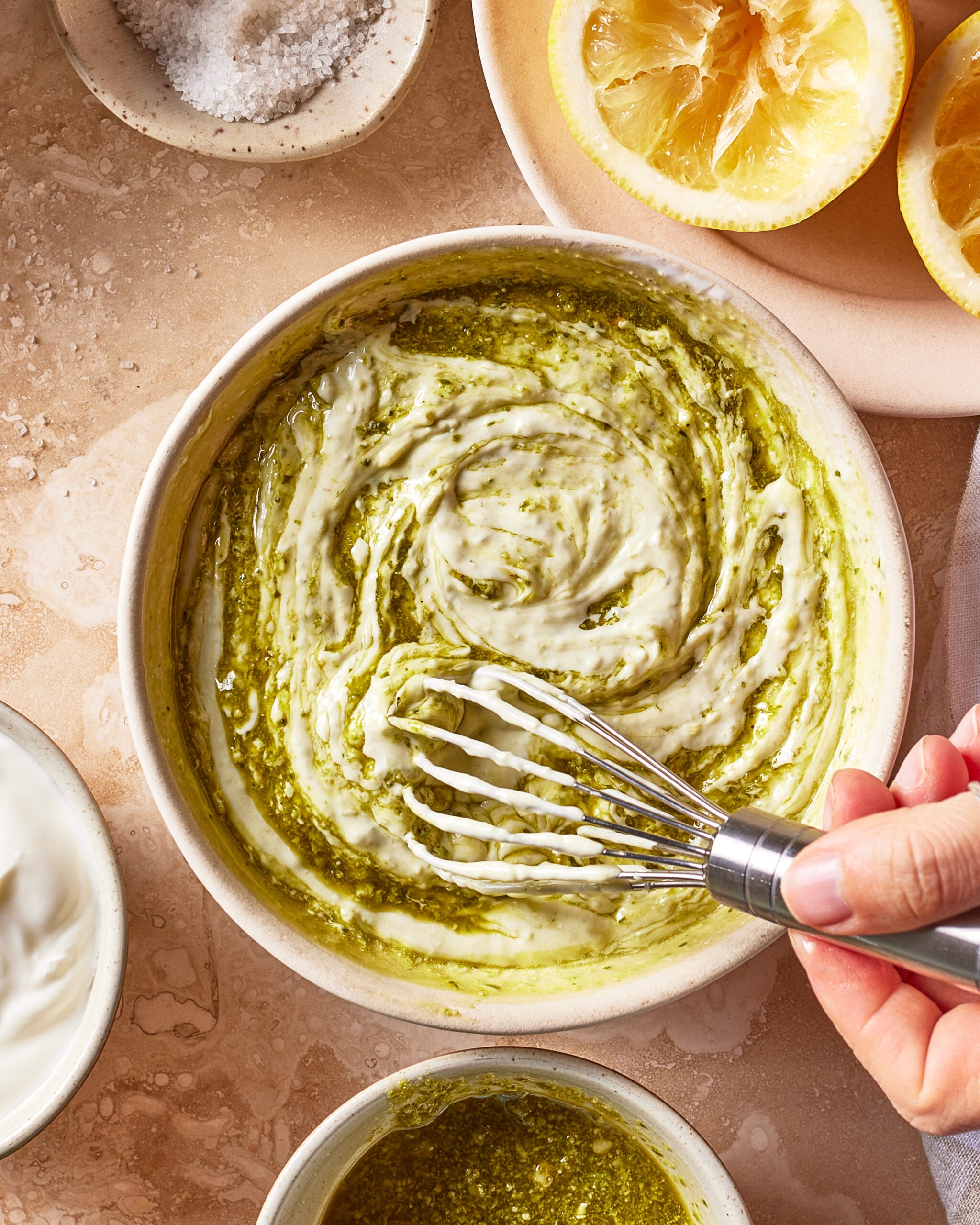 A hand uses a whisk to mix a green herb sauce into a creamy white base in a bowl, surrounded by lemon halves, salt, and small bowls of ingredients on a beige surface.