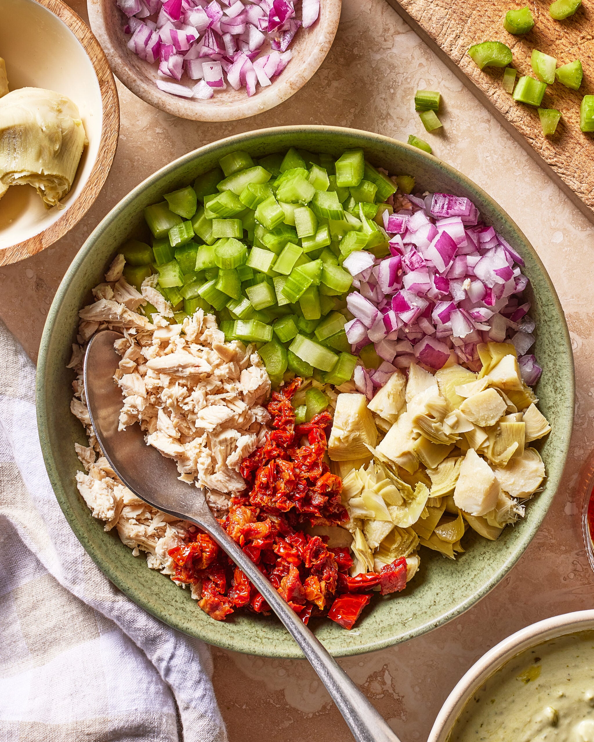 A bowl with chopped celery, red onion, sun-dried tomato, artichoke hearts, and shredded chicken, with a spoon inside. Other vegetables and a creamy dressing are nearby on a countertop.