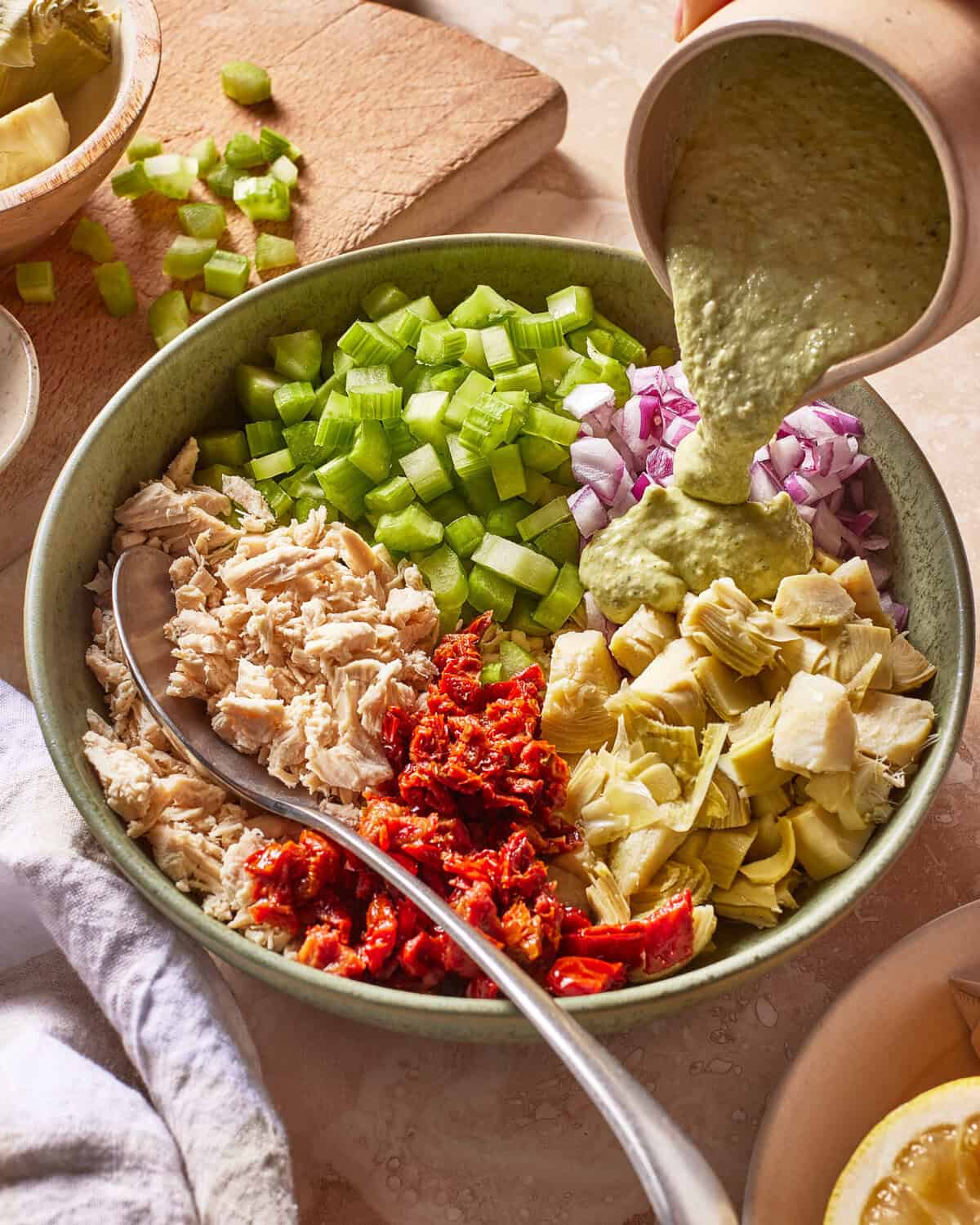 A bowl with shredded chicken, chopped celery, red onion, sun-dried tomatoes, and artichoke hearts, with green dressing being poured on top. A spoon rests in the bowl and extra celery is on a cutting board nearby.