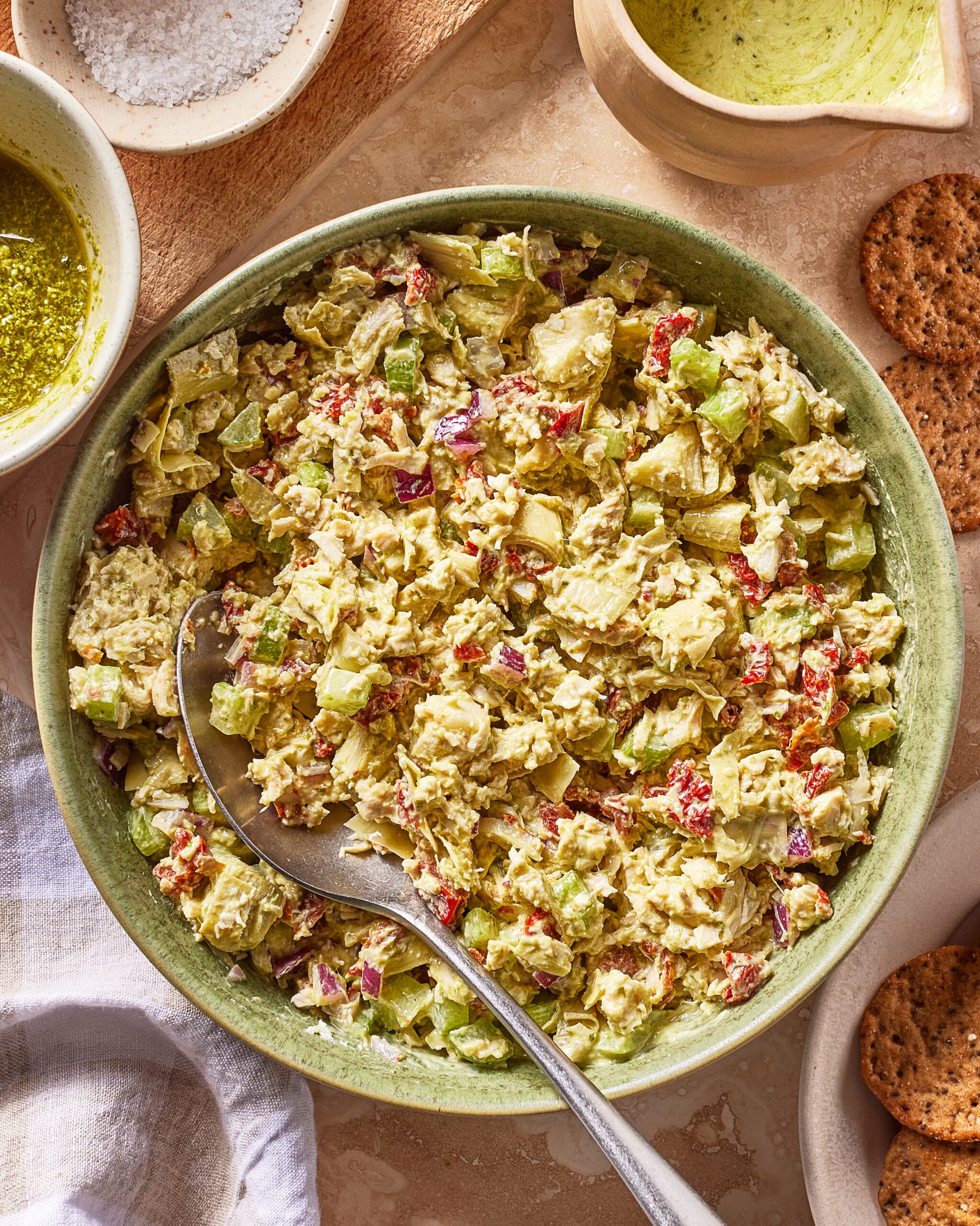 A green bowl filled with a chunky salad containing chopped vegetables and herbs, with a large serving spoon. Surrounding items include crackers, a cloth, sauces, and a dish of salt.