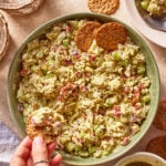 A hand dips a round cracker into a bowl of chunky vegetable salad with chopped celery, red onion, and herbs. Crackers and a small plate of salad are nearby on a beige surface.