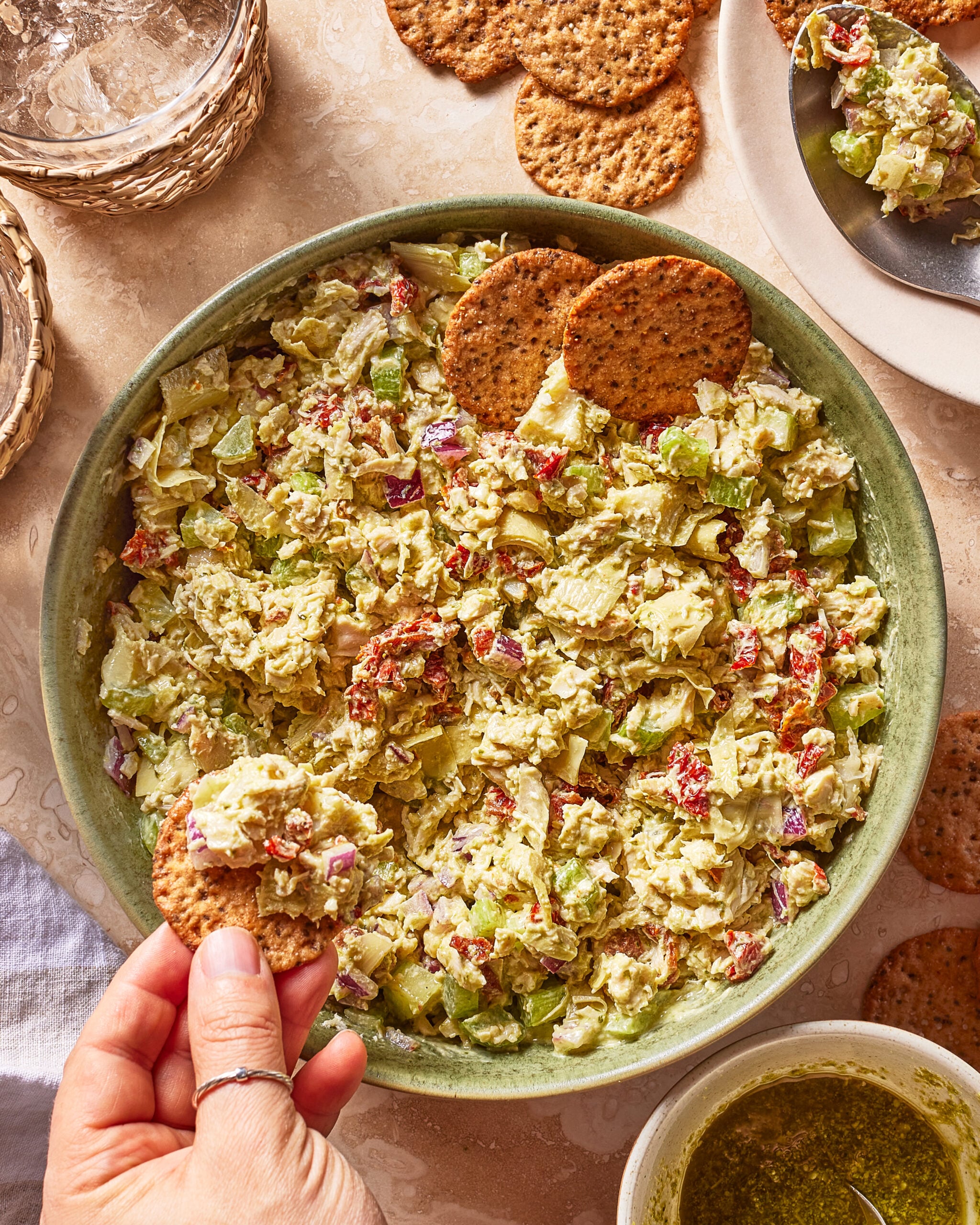 A hand dips a round cracker into a bowl of chunky vegetable salad with chopped celery, red onion, and herbs. Crackers and a small plate of salad are nearby on a beige surface.