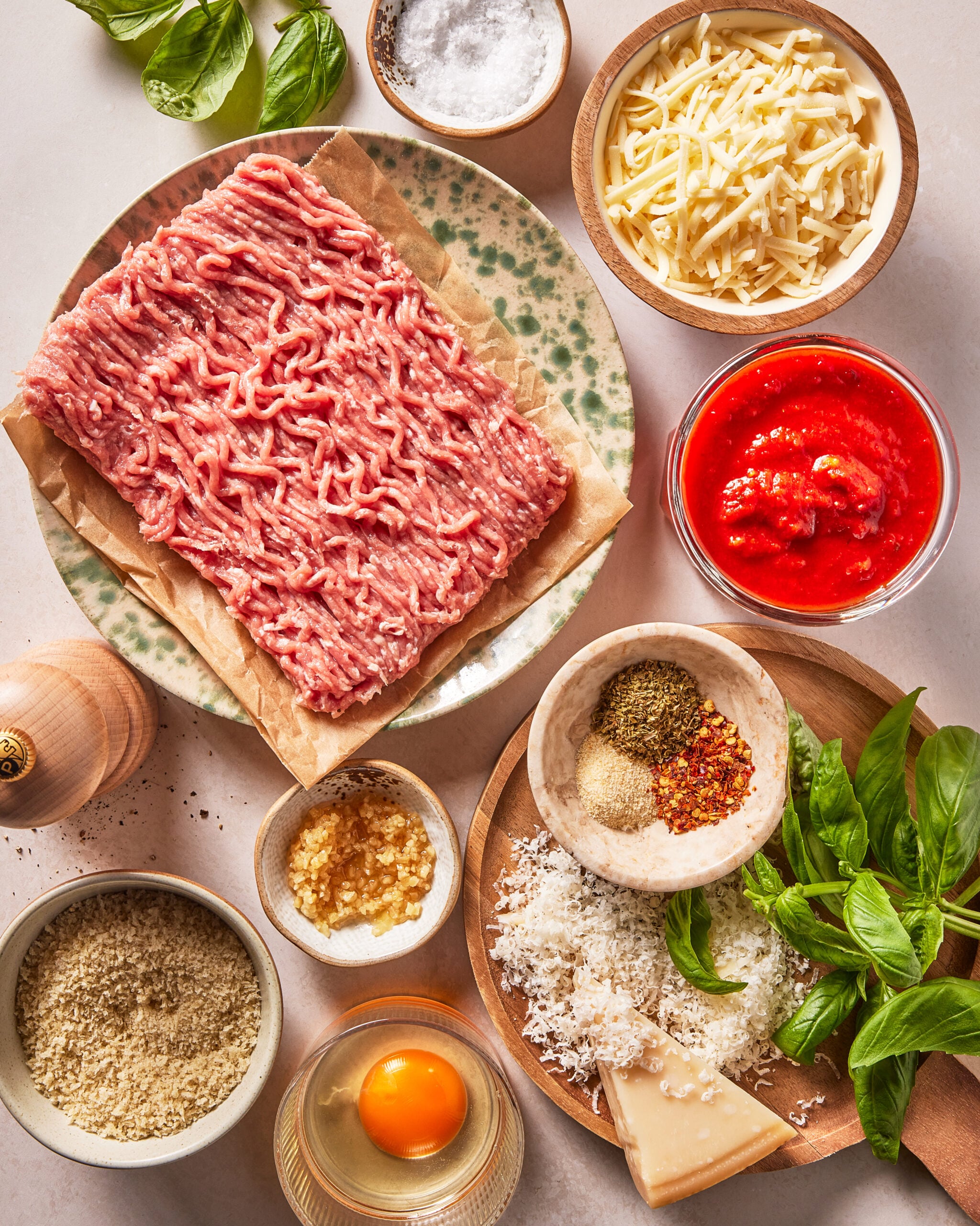 Ground meat on a plate, surrounded by grated cheese, tomato sauce, spices, basil, an egg, breadcrumbs, garlic, pepper, and salt, arranged neatly on a light tabletop.