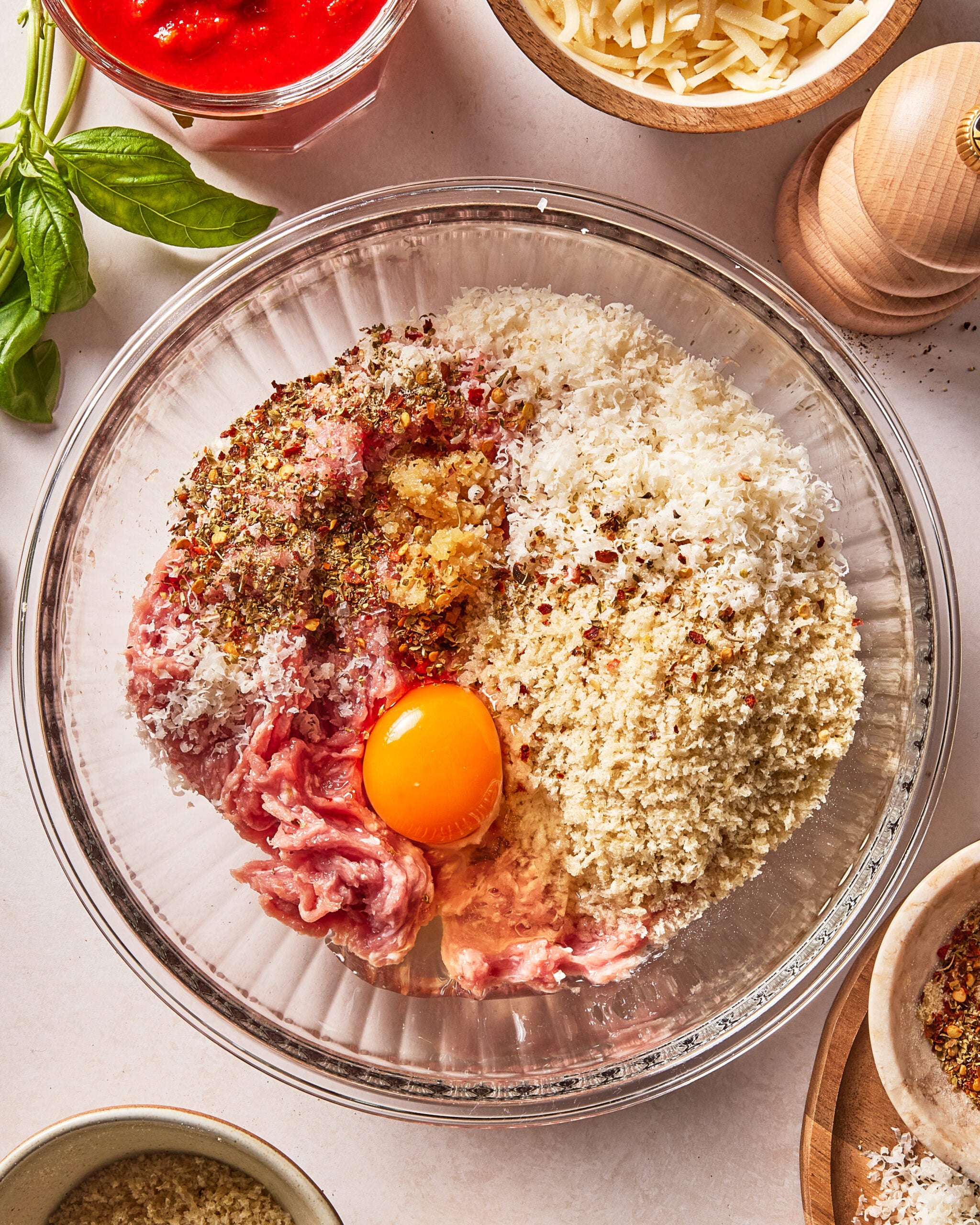 A glass bowl with ground meat, an egg, breadcrumbs, grated cheese, and seasonings, surrounded by bowls of tomato sauce, cheese, basil, and a pepper grinder on a light countertop.