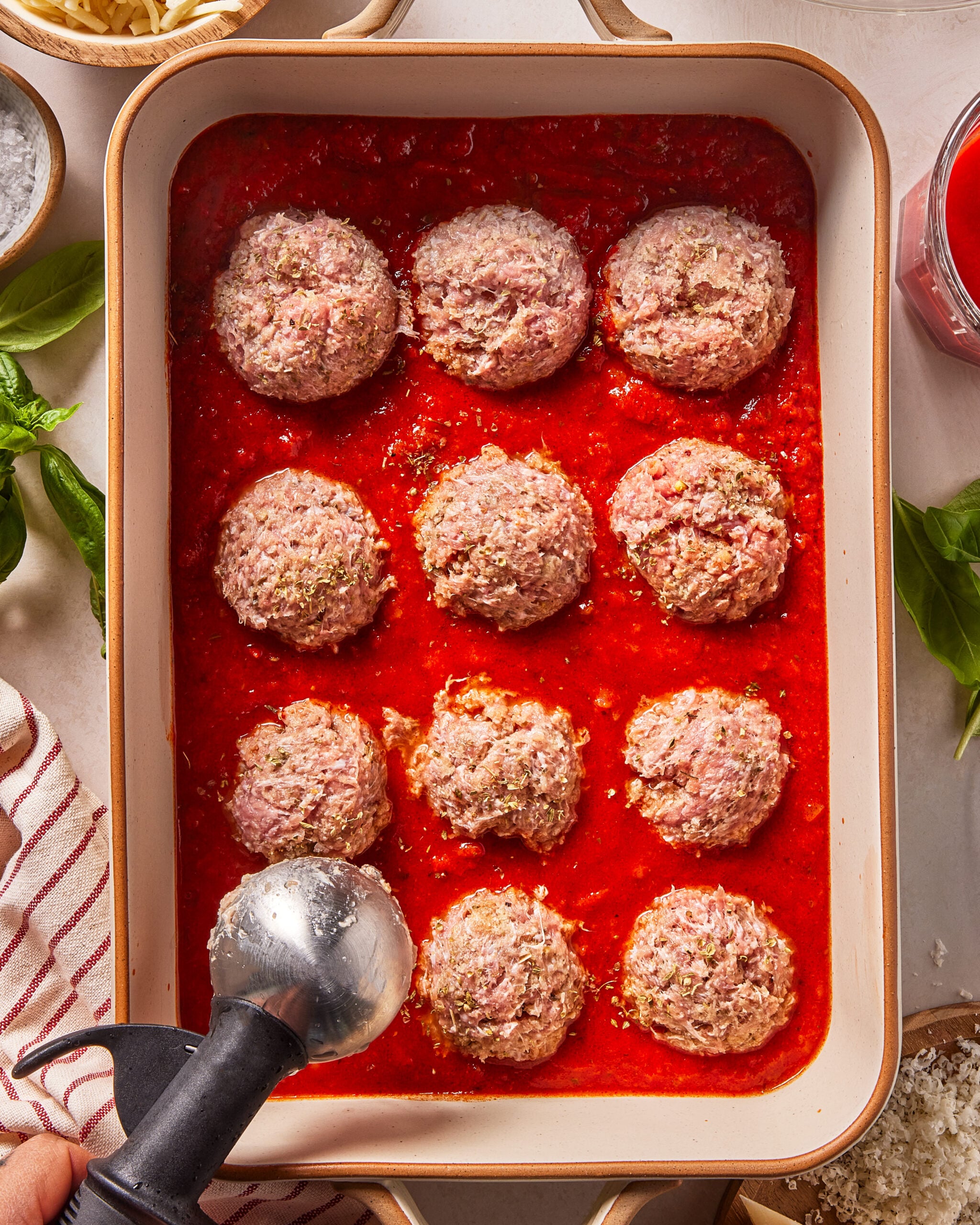 A baking dish with 12 raw meatballs in red tomato sauce. An ice cream scoop is placing a meatball in the pan. Fresh herbs, grated cheese, and a striped towel are nearby.