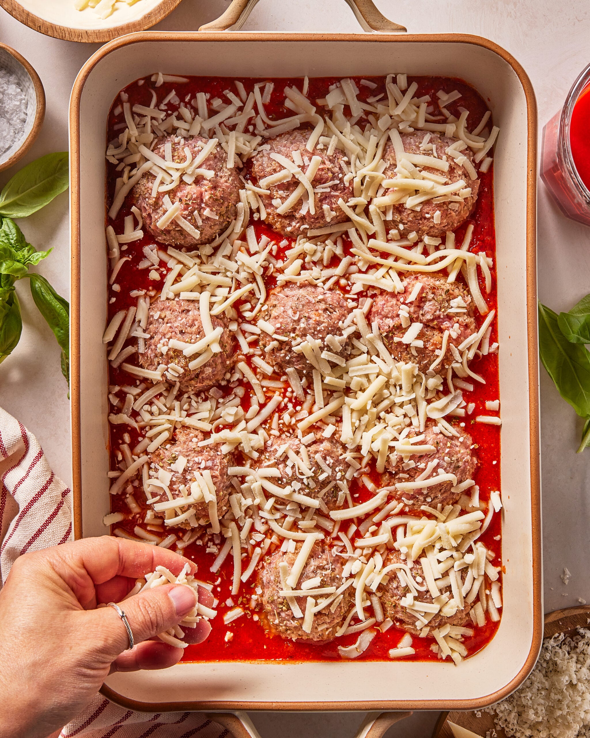 A hand sprinkles shredded cheese over raw meatballs in tomato sauce in a rectangular baking dish, surrounded by bowls of ingredients and fresh basil leaves on a white surface.