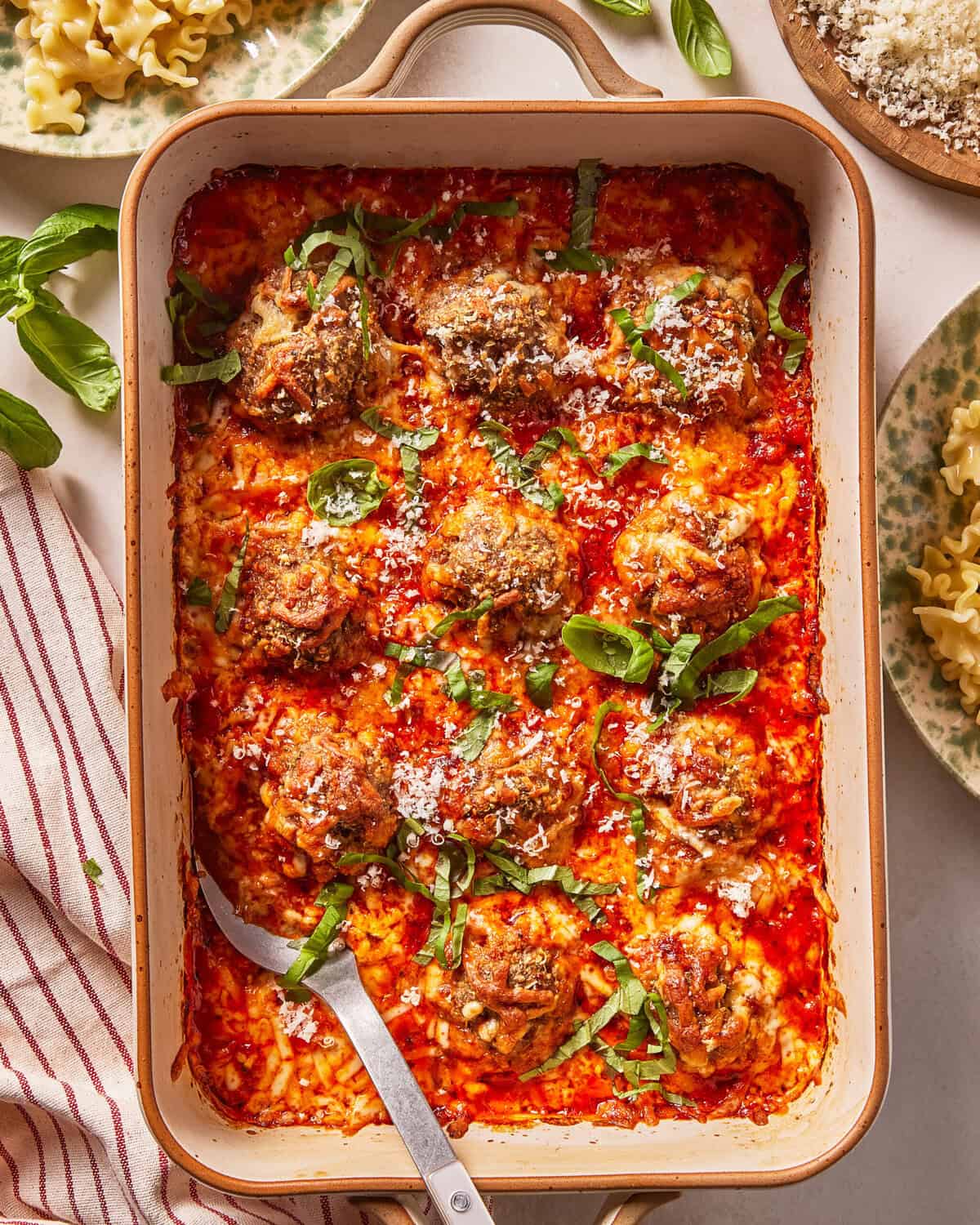 A baking dish filled with meatballs in tomato sauce, topped with melted cheese and fresh basil, with a serving spoon. Plate of pasta and grated cheese are nearby.