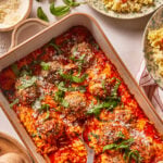 A casserole dish of baked lasagna topped with meatballs, tomato sauce, fresh basil, and grated cheese, with a serving spoon. Plates of pasta and bowls of grated cheese are nearby on a white table.