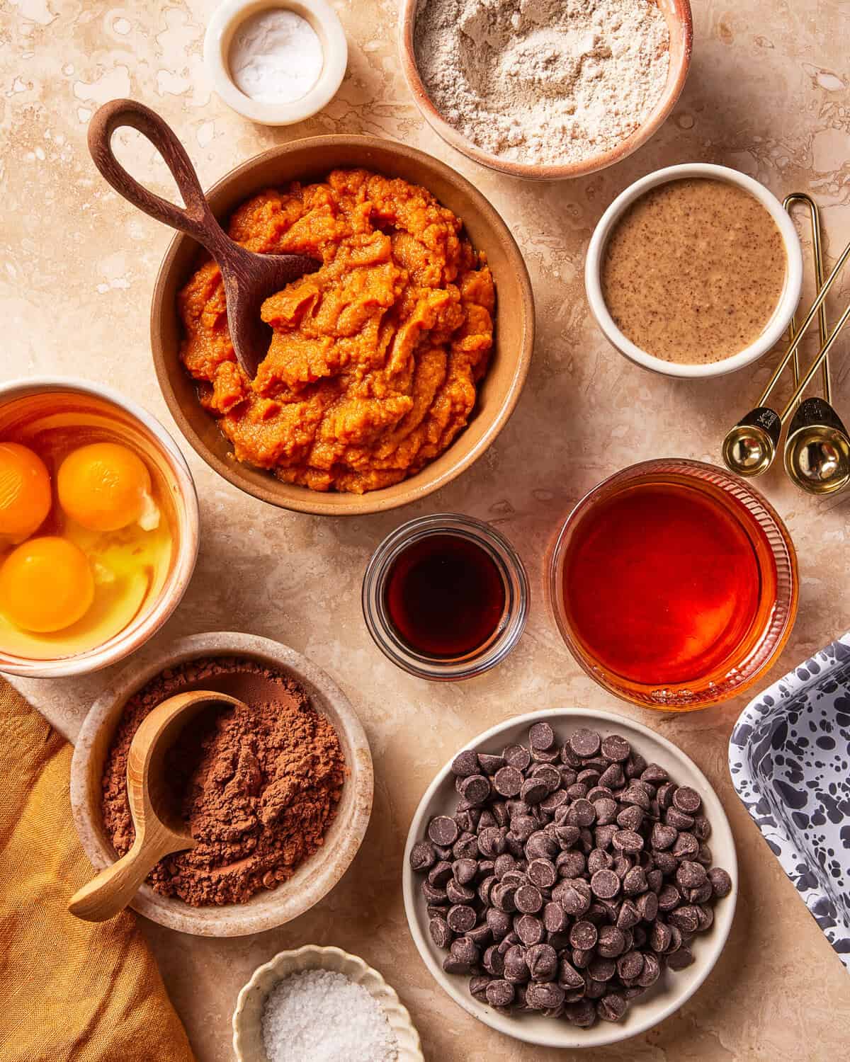 Various baking ingredients are arranged on a countertop, including pumpkin puree, eggs, flour, cocoa powder, chocolate chips, vanilla, oil, nut butter, salt, and measuring spoons.
