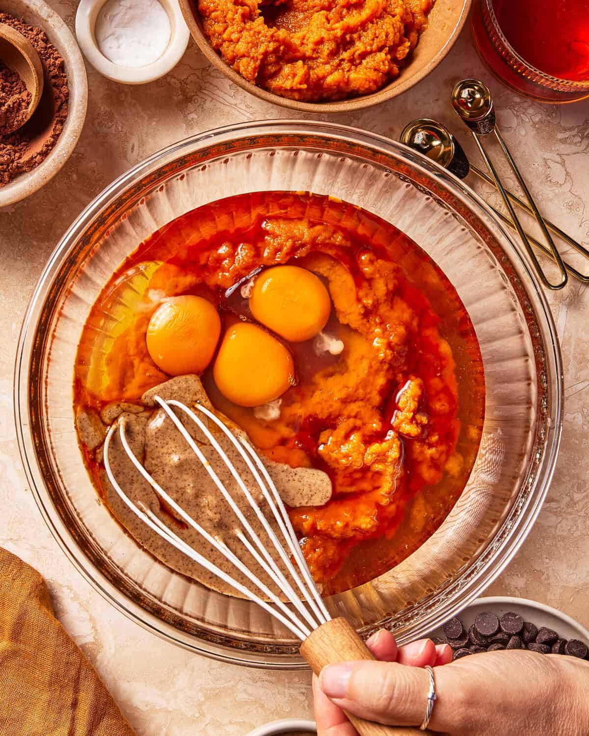 A hand holds a whisk in a glass bowl with eggs, pumpkin puree, and wet ingredients, surrounded by chocolate chips, measuring spoons, and baking supplies on a countertop.