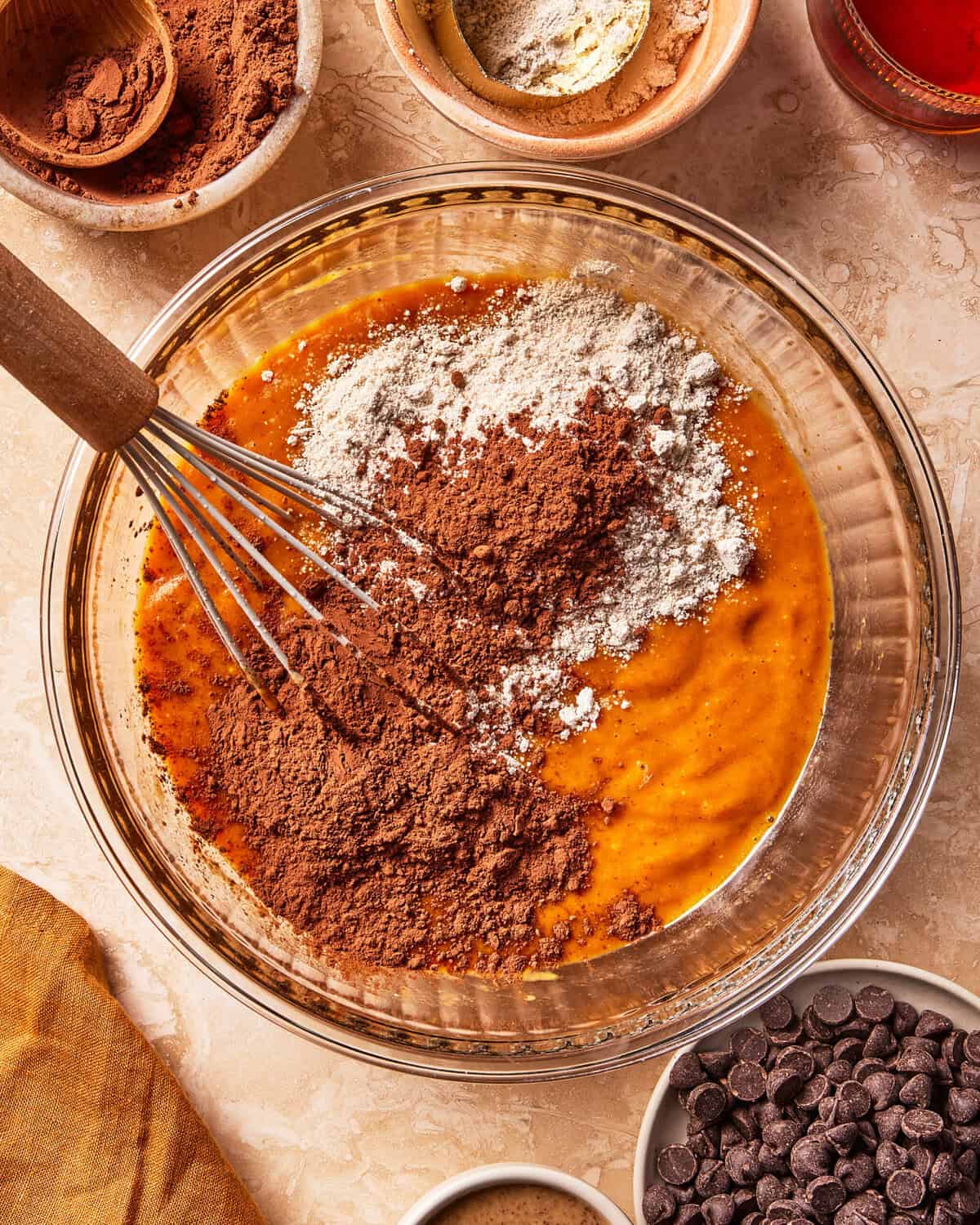 A glass bowl with cocoa powder, flour, and wet ingredients being whisked together, surrounded by bowls of chocolate chips, cocoa, and flour on a beige countertop.