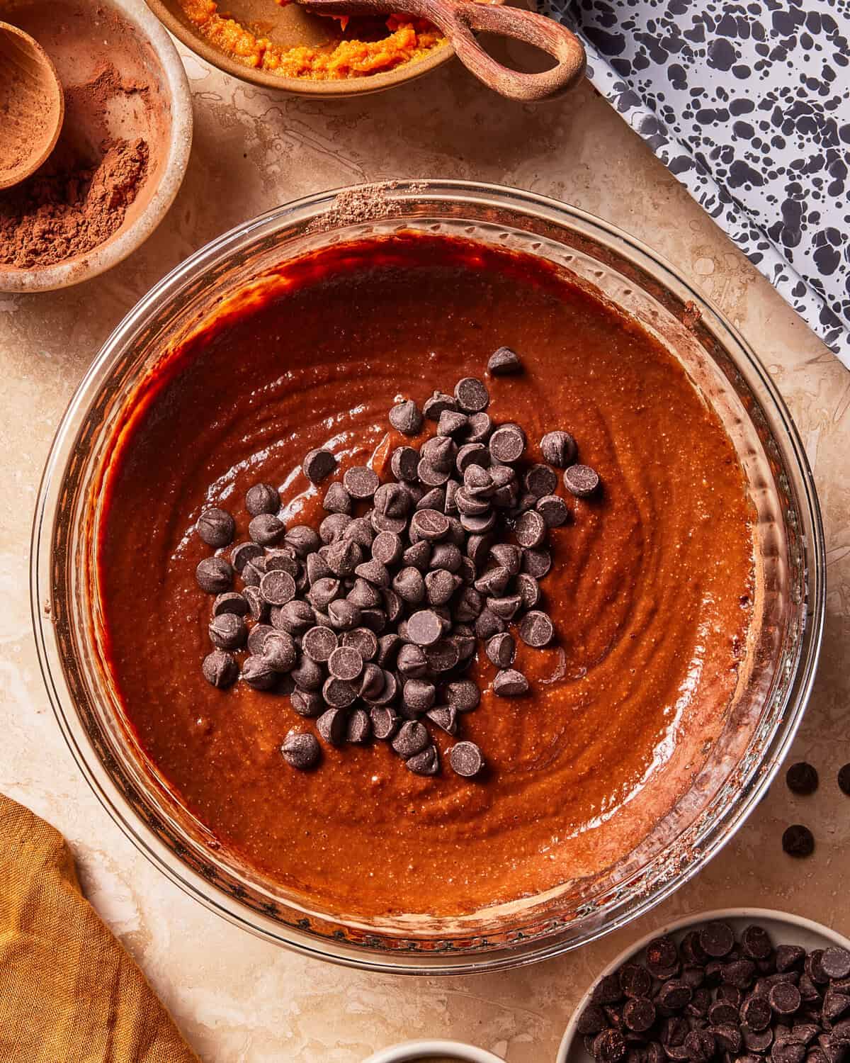 A glass bowl filled with brownie batter topped with chocolate chips sits on a counter, surrounded by small bowls of ingredients and a yellow cloth.