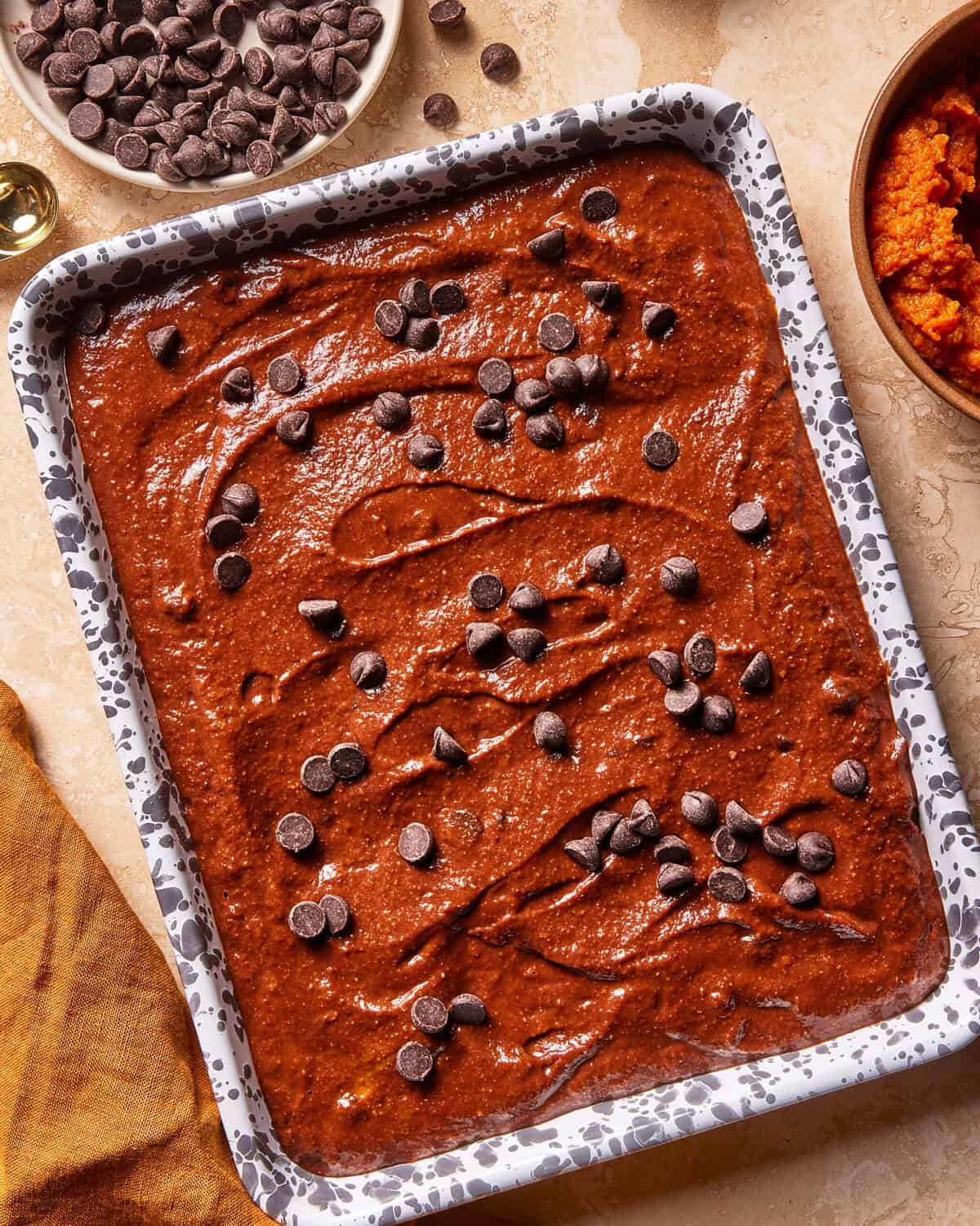 A speckled baking dish filled with chocolate brownie batter, topped with chocolate chips, sits on a countertop beside bowls of chocolate chips and pumpkin puree.