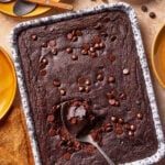 A tray of chocolate brownies with chocolate chips on top, one brownie scooped out with a spoon. Surrounding the tray are yellow plates, utensils, a bowl of chocolate chips, and a napkin.