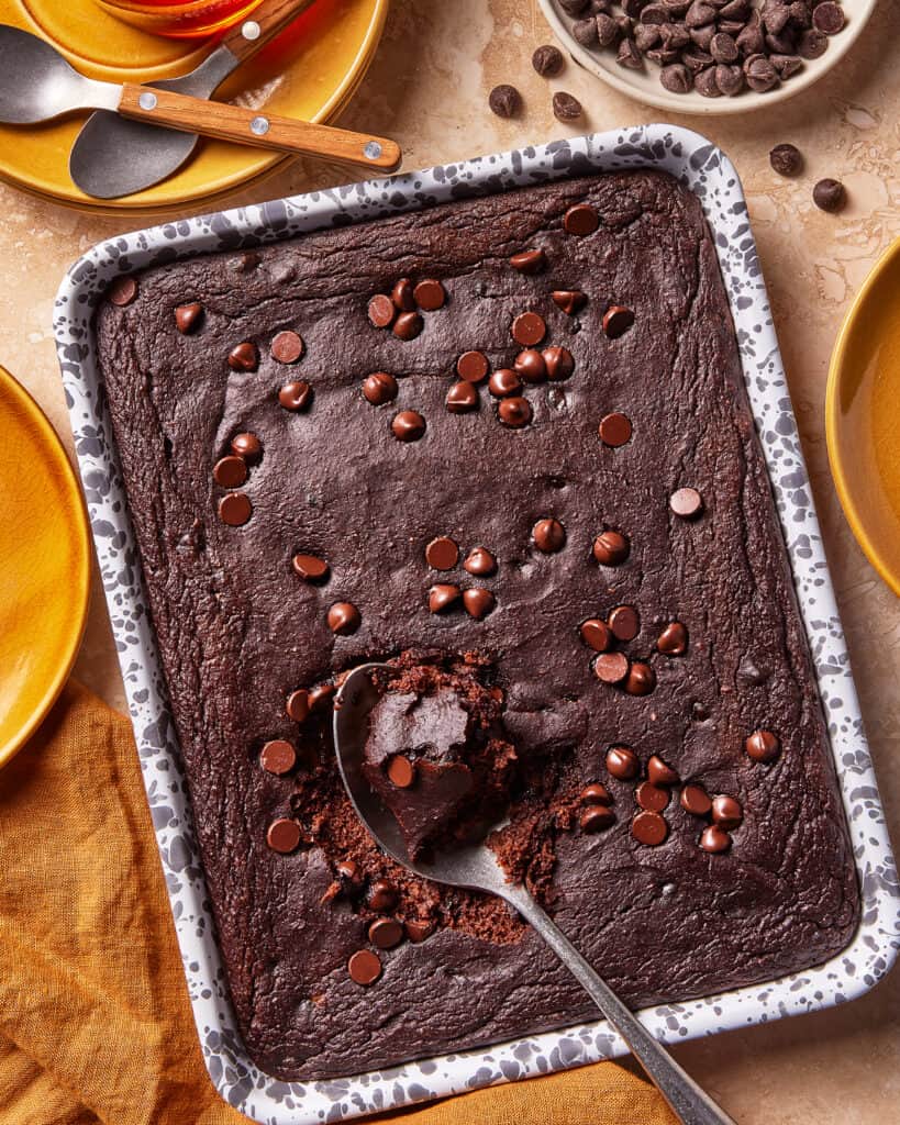 A tray of chocolate brownies with chocolate chips on top, one brownie scooped out with a spoon. Surrounding the tray are yellow plates, utensils, a bowl of chocolate chips, and a napkin.