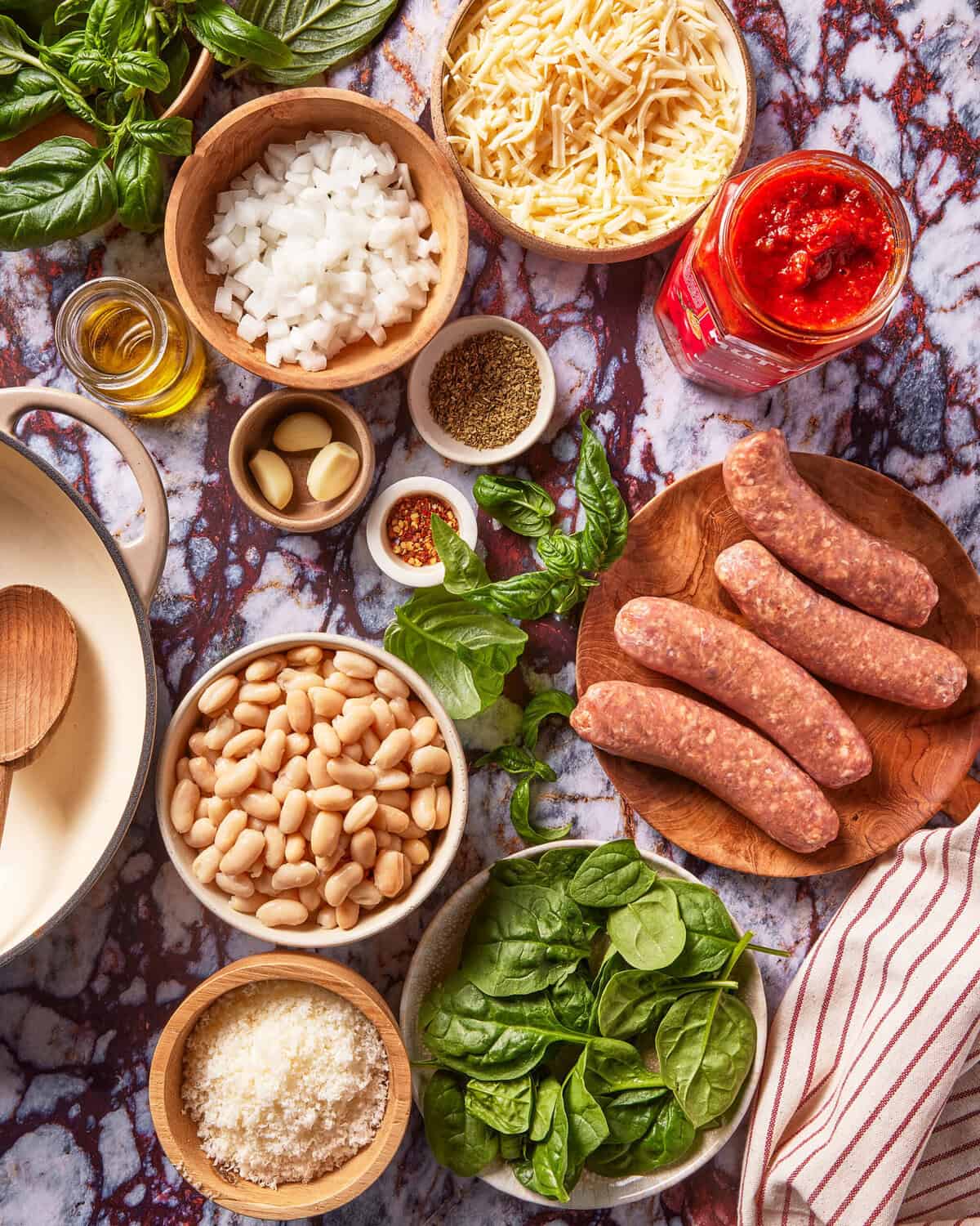 A top-down view of ingredients on a marble surface, including sausage, beans, spinach, cheese, garlic, chopped onion, tomato sauce, spices, olive oil, and fresh basil, ready for cooking.