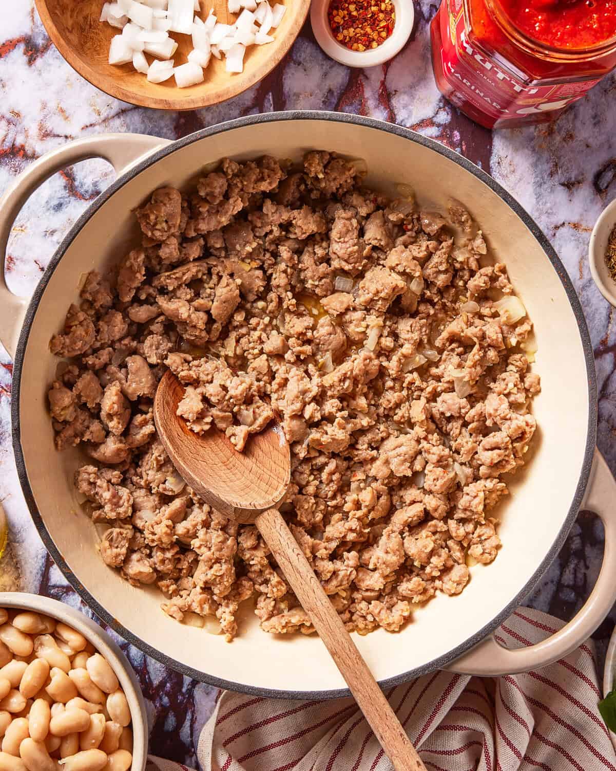 A pot of browned ground meat with a wooden spoon, surrounded by bowls of chopped onions, beans, red pepper flakes, and a jar of red sauce on a marble surface.
