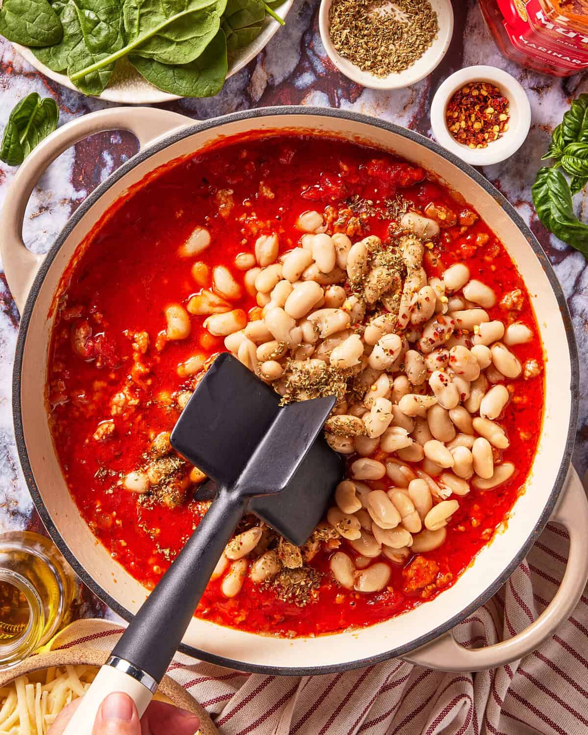 A pot of tomato-based stew with white beans, Italian herbs, and a black spatula, surrounded by fresh spinach, olive oil, dried herbs, and basil on a marble countertop.