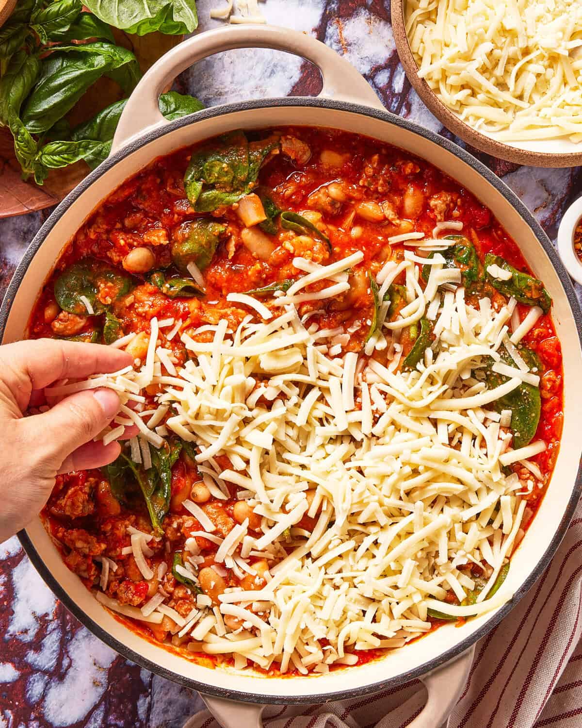 A hand sprinkles shredded cheese over a pan filled with tomato sauce, beans, and spinach. Fresh basil and more cheese are in bowls nearby on a marble countertop.