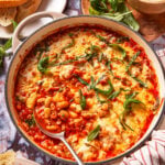 A casserole dish filled with baked beans in tomato sauce, topped with melted cheese and fresh basil leaves. Slices of bread, basil, grated cheese, and seasonings are arranged around the dish.
