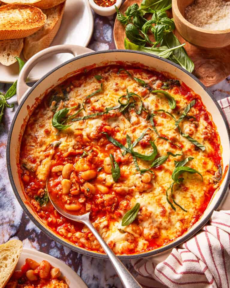 A casserole dish filled with baked beans in tomato sauce, topped with melted cheese and fresh basil leaves. Slices of bread, basil, grated cheese, and seasonings are arranged around the dish.