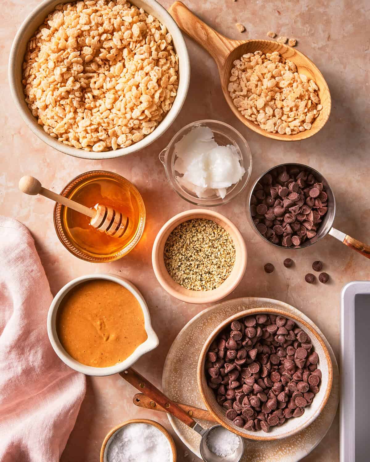 Bowls and cups filled with Rice Krispies cereal, chocolate chips, nut butter, honey, coconut oil, hemp seeds, and salt are arranged on a beige countertop with a pink cloth.