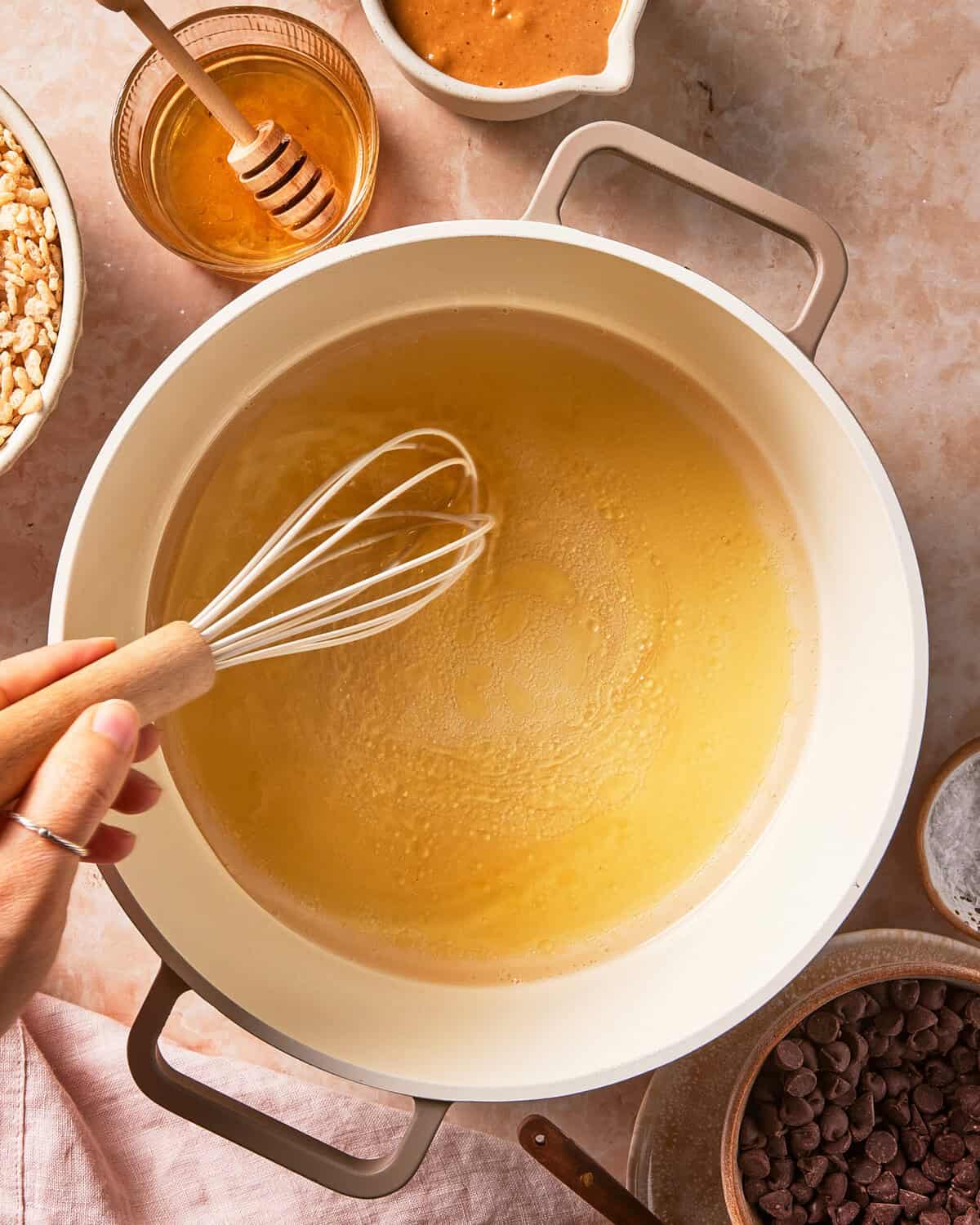 A hand whisks a yellow liquid in a white pot, surrounded by bowls of honey, chocolate chips, peanut butter, and puffed rice on a light countertop.