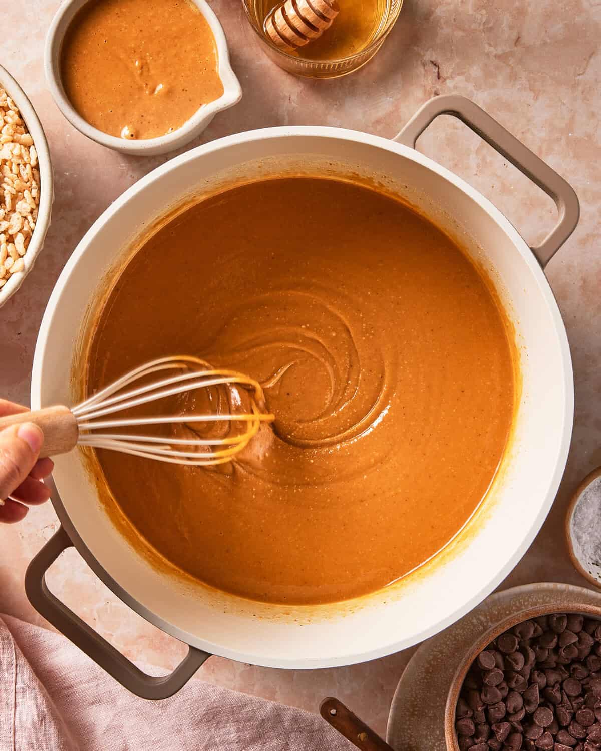 A hand whisks a creamy, caramel-colored mixture in a white pot, surrounded by bowls of chocolate chips, puffed rice, peanut butter, and honey on a light countertop.