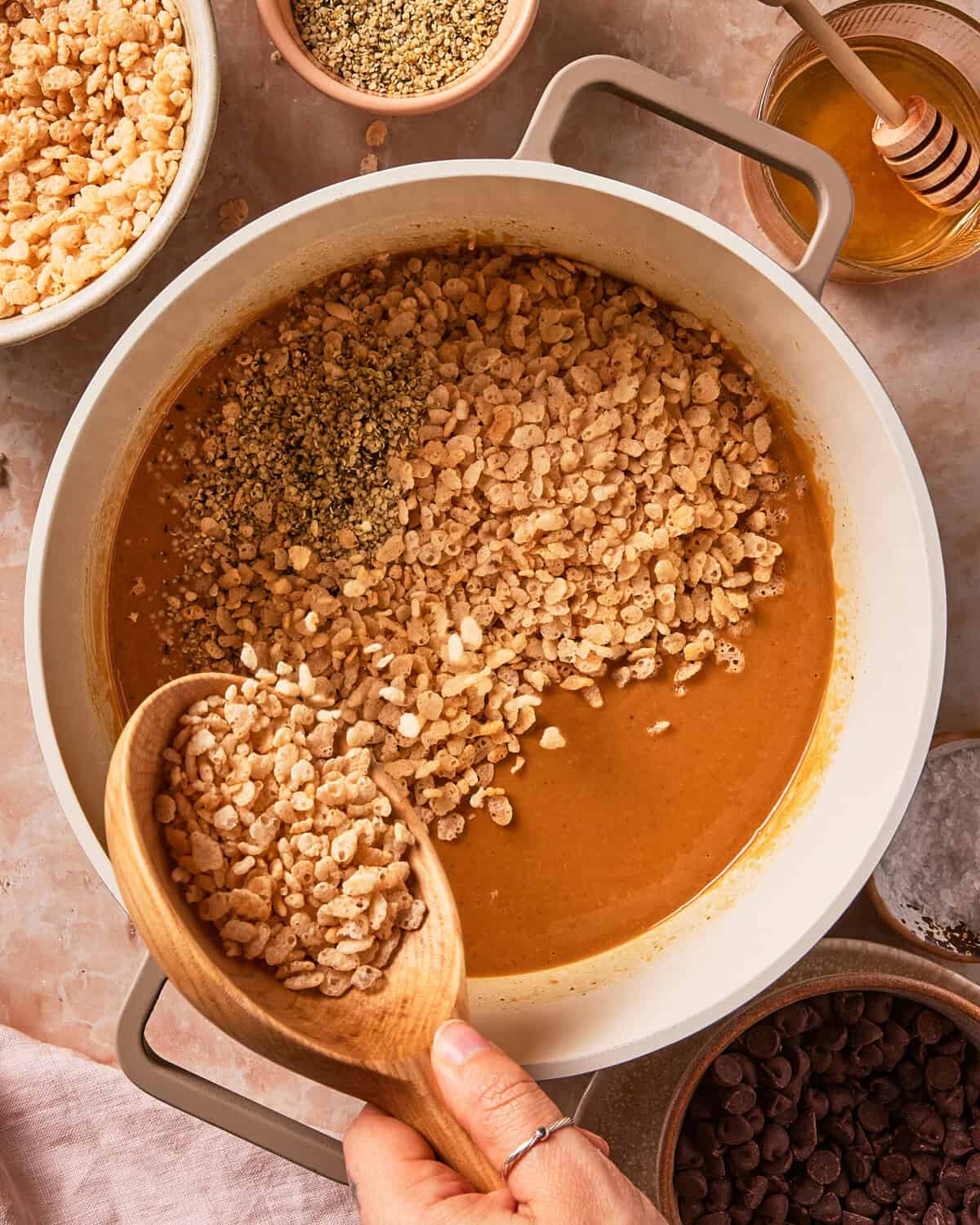 A hand adds crispy rice cereal to a pot with a peanut butter mixture. Surrounding the pot are bowls of crispy rice, seeds, honey with a dipper, and chocolate chips.