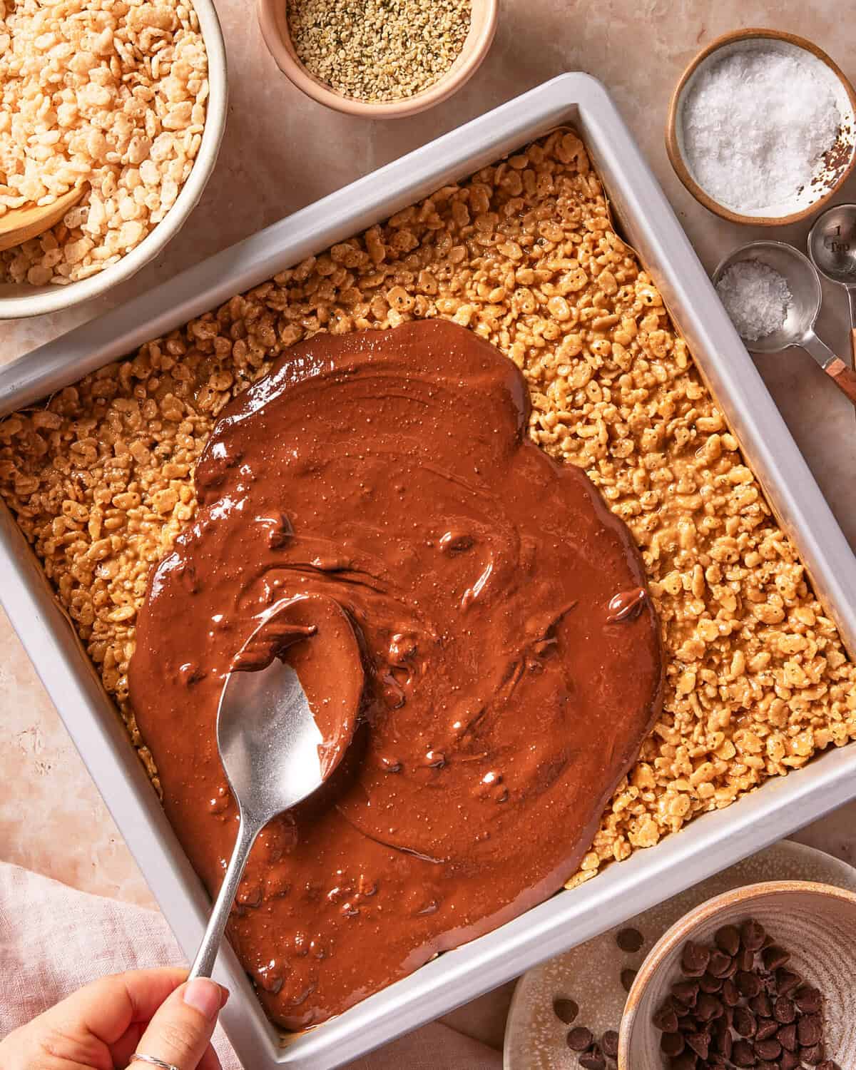 A hand spreads chocolate over Rice Krispies treats in a baking pan, surrounded by bowls of rice cereal, chocolate chips, hemp seeds, and salt on a light countertop.
