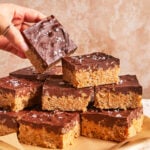 A hand picks up a chocolate-topped dessert bar from a plate stacked with similar bars. The bars have a crispy base and are sprinkled with flaky sea salt.